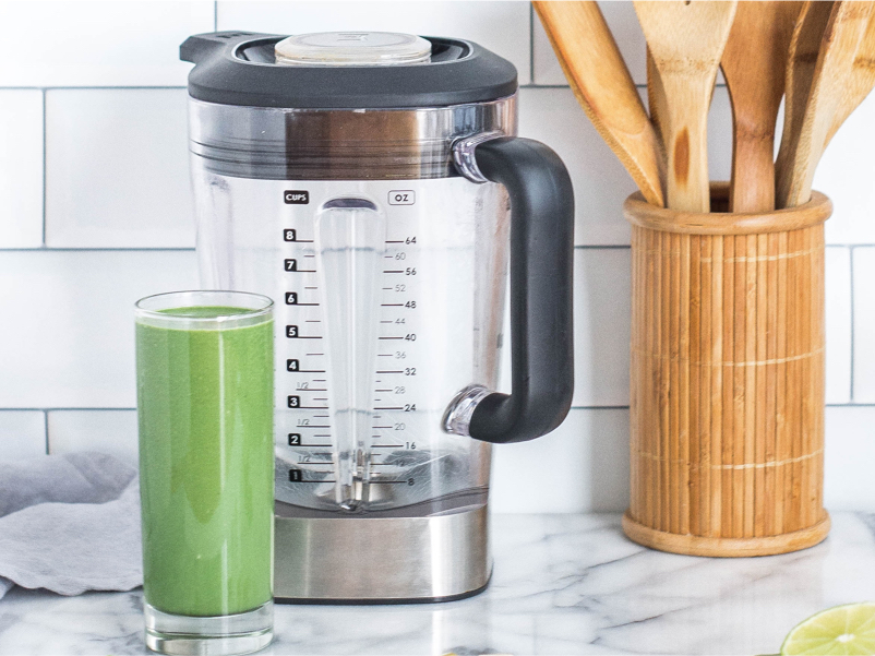 An empty blender with a cup full of green smoothie in front of it